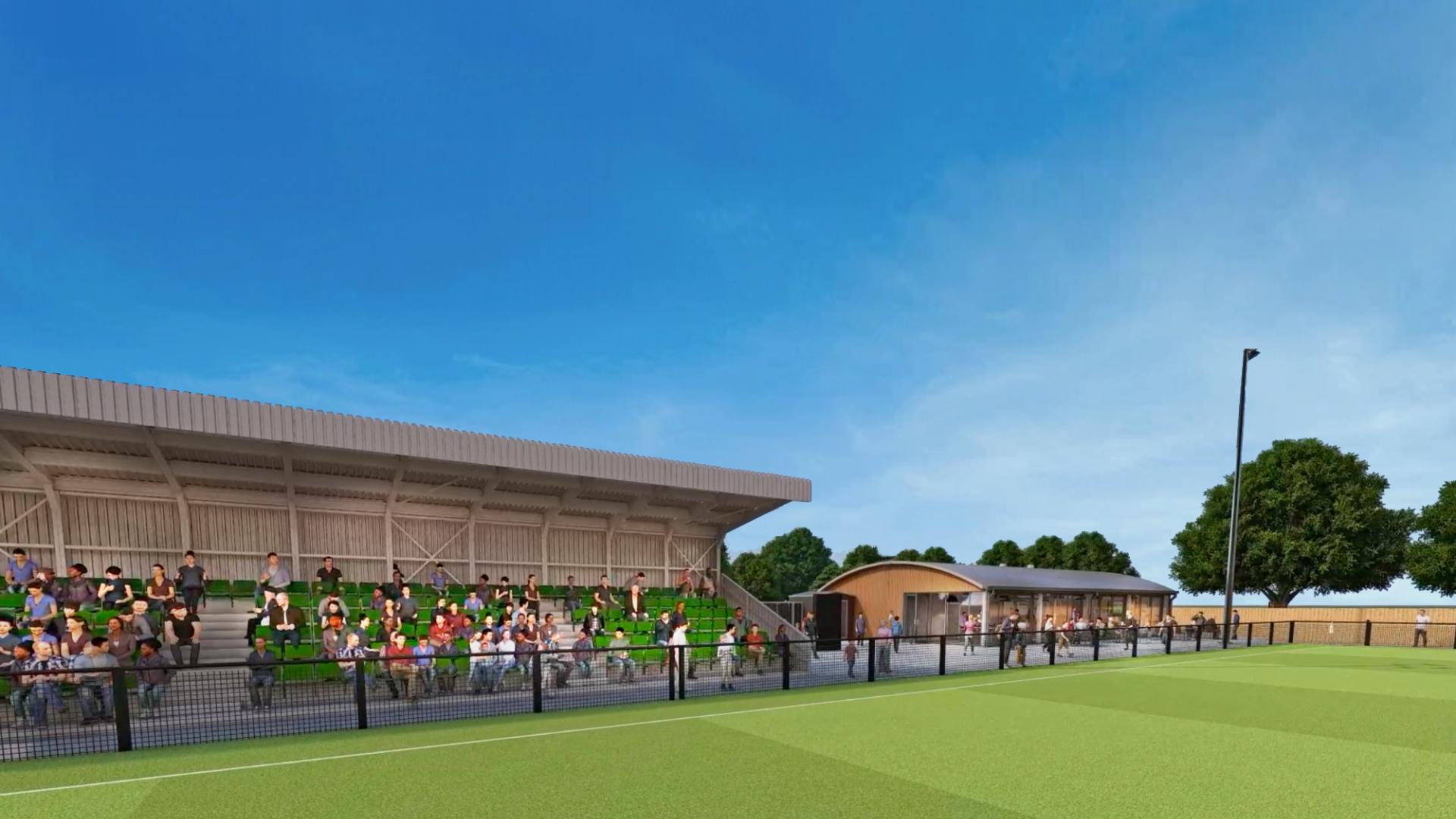 The image depicts a vibrant outdoor sports facility under a clear blue sky. A large, modern grandstand with a metal roof accommodates a diverse crowd of spectators dressed in casual attire. Adjacent to the grandstand is a sprawling, immaculate green turf field, delineated for athletic activities. In the background, there are well-designed, contemporary structures, likely facilities or concessions, enhancing the venue's community-friendly atmosphere